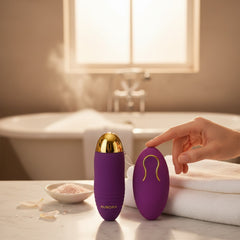 Bala vibradora morada de silicón Glow&Vibe con control remoto, diseño ergonómico y detalles dorados en baño de lujo.