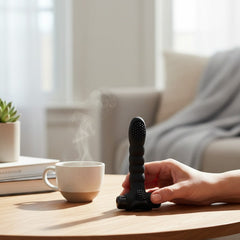 Dedal vibrador negro Glow&Vibe Love Alexander de silicón texturizado sobre mesa de madera junto a una taza de café